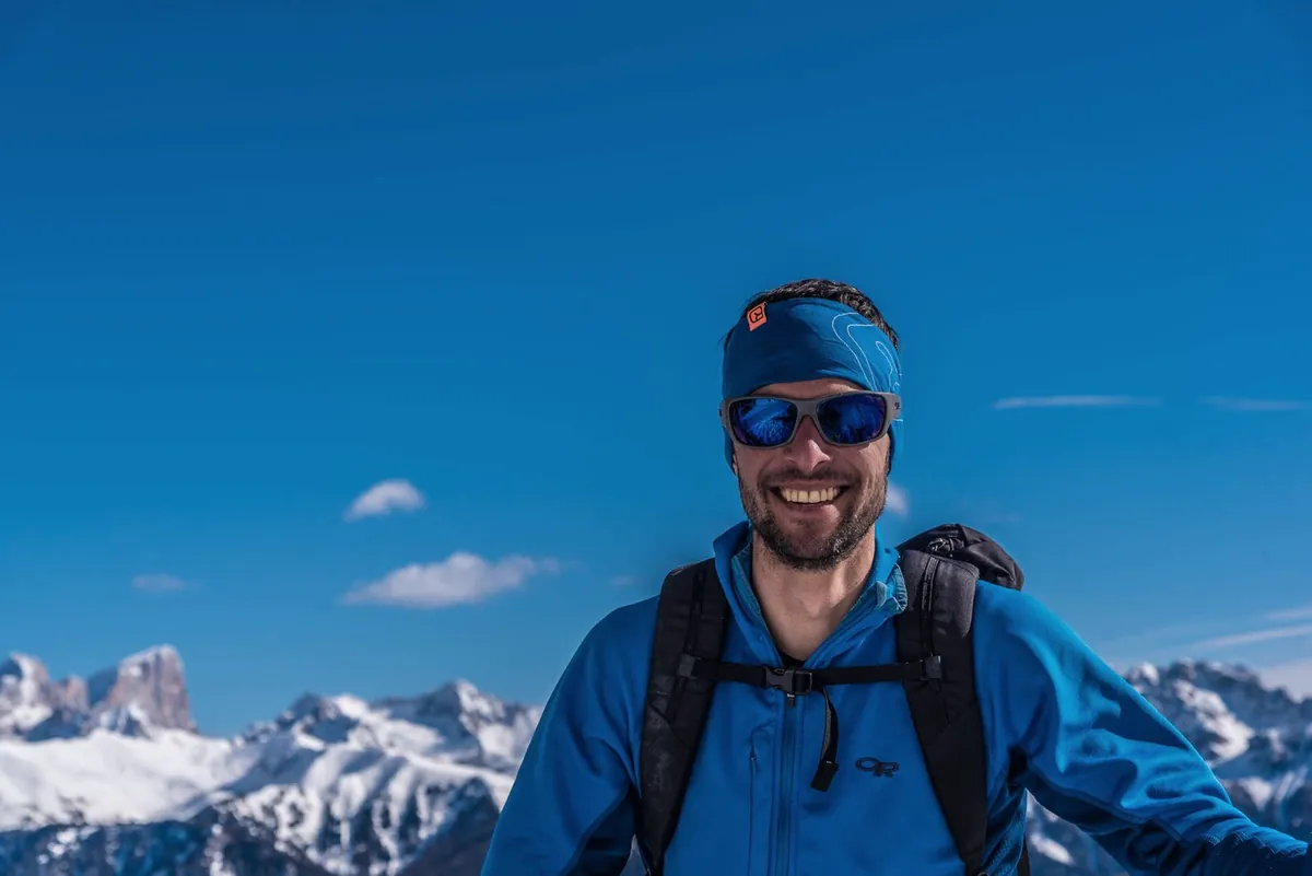 Thomas Engl lächelnd mit Stirnband und Rucksack vor verschneiten Gipfeln